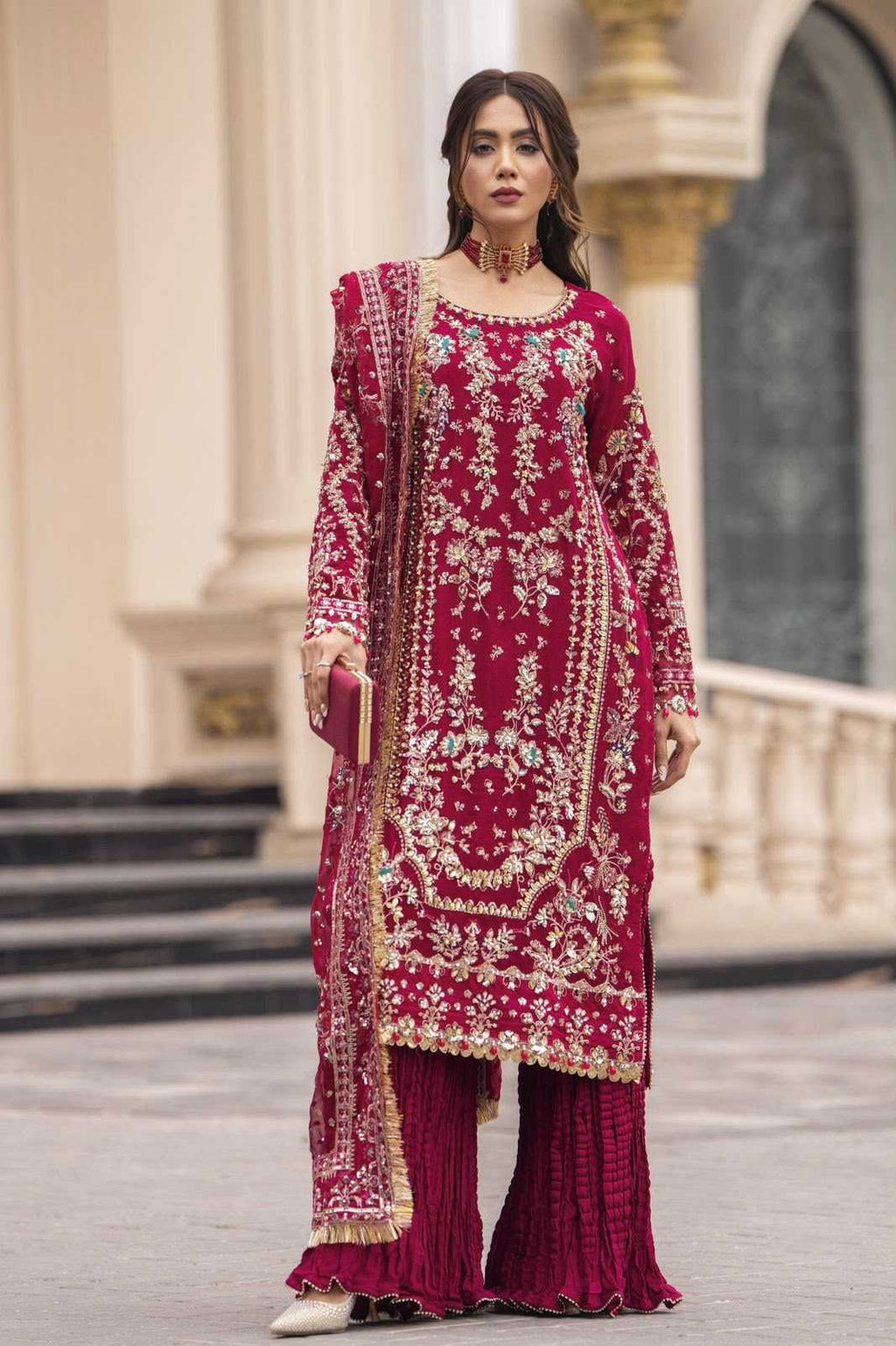 A woman modeling a traditional deep magenta three-piece salwar kameez suit with intricate gold embroidery, pleated matching pants, and a coordinating dupatta, set against a classical stone building background.