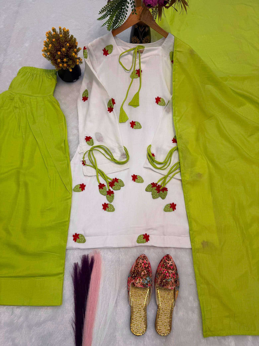 A women's traditional outfit laid flat on a white textured surface, featuring a white kurta with red and green floral embroidery and tie details, paired with bright lime green pants and a matching lime green scarf. A pair of floral embroidered flat shoes is also placed below.