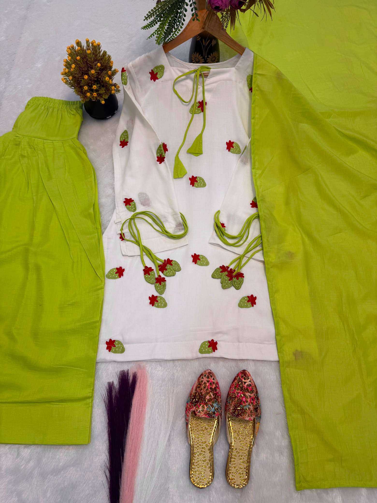 A women's traditional outfit laid flat on a white textured surface, featuring a white kurta with red and green floral embroidery and tie details, paired with bright lime green pants and a matching lime green scarf. A pair of floral embroidered flat shoes is also placed below.
