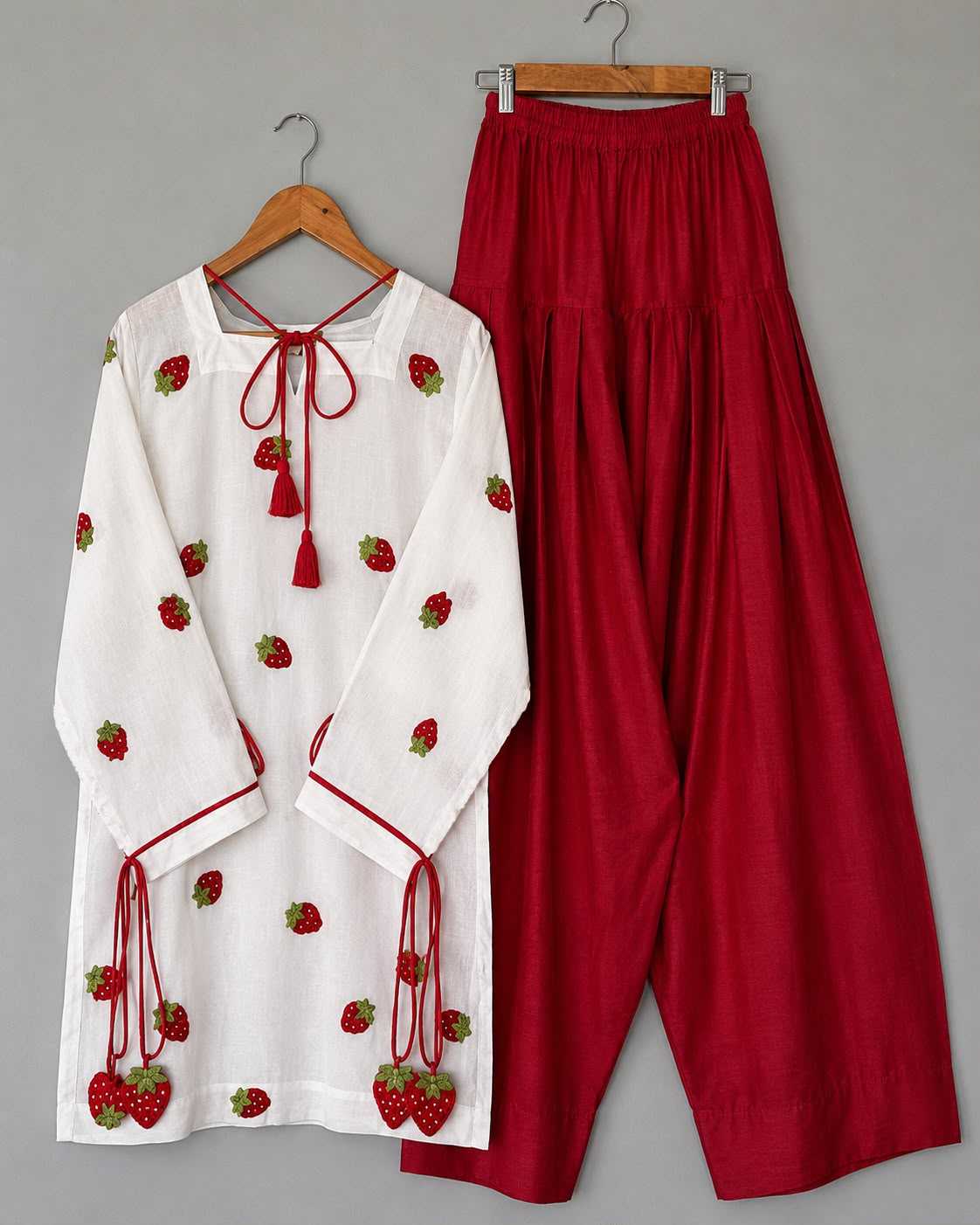 A two-piece women's outfit consisting of a white long-sleeve tunic top with strawberry embroidery and red drawstrings, paired with high-waisted red wide-leg pants, displayed on wooden hangers against a neutral gray background.