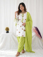 A woman models a white salwar kameez suit with green embroidery, matching green pants, and a lime green dupatta, set against a white draped background with decorative dried grass.
