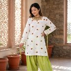 A woman models a white long-sleeve kurta decorated with small red and green strawberry motifs, paired with lime green pants, standing in a sunlit courtyard with stone walls and potted plants.