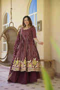 A woman models a deep maroon Anarkali gown with vertical white stripes and a colorful floral border, set in a warm, sunlit courtyard with a hanging wicker chair.