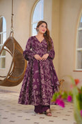 Woman in a purple floral dress standing in a room with a wicker chair and decorative plants.
