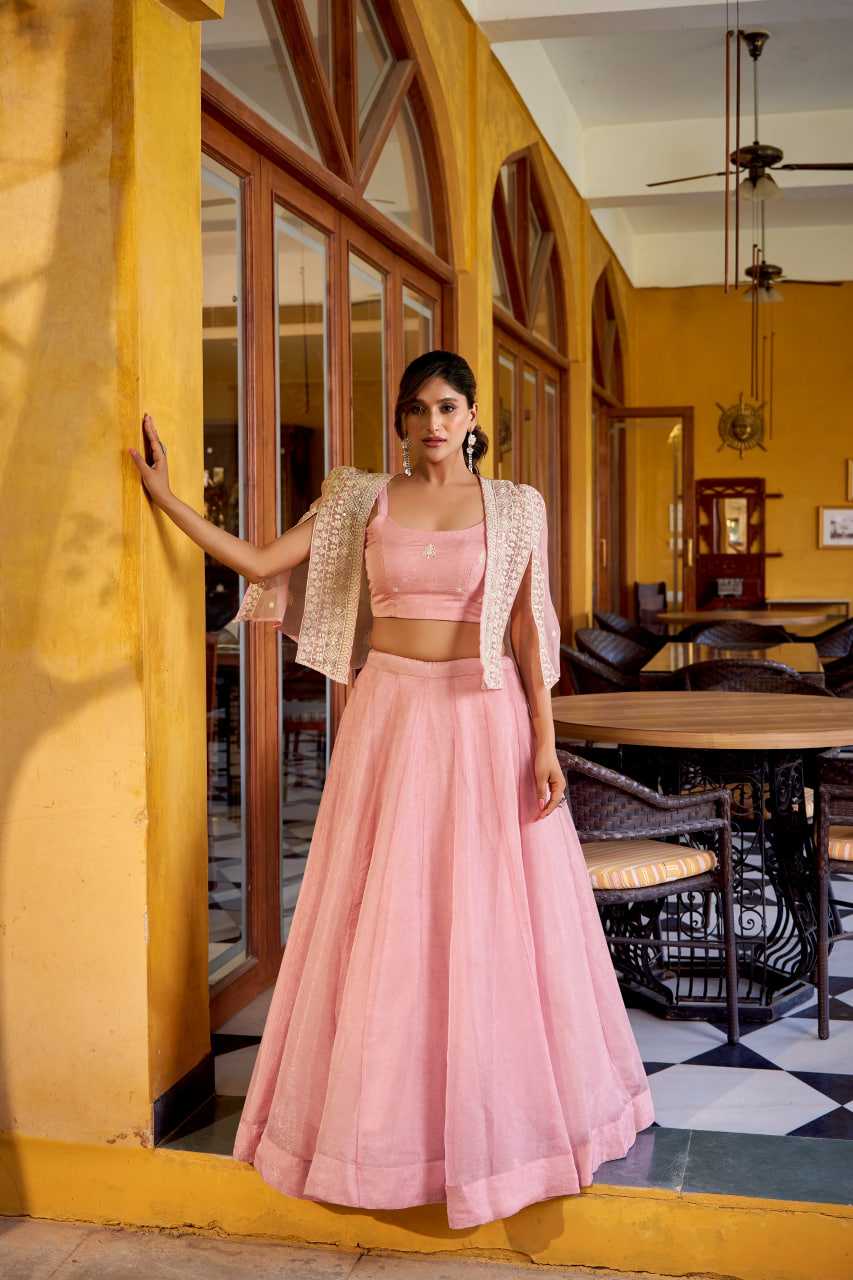 A woman models a light pink two-piece traditional outfit with a matching flared skirt, crop top, and a short beige embroidered jacket, standing in a warm yellow-toned indoor cafe setting.