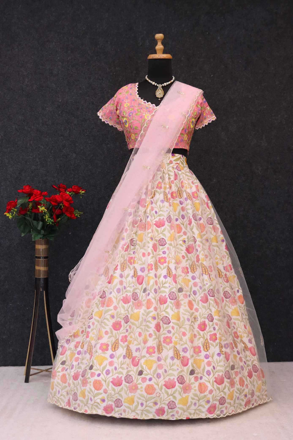 A pink and cream floral embroidered lehenga choli set displayed on a mannequin. The set features a pink embroidered blouse with short sleeves, a matching pleated skirt decorated with pink, yellow, and orange floral patterns, and a sheer light pink dupatta draped over the skirt.