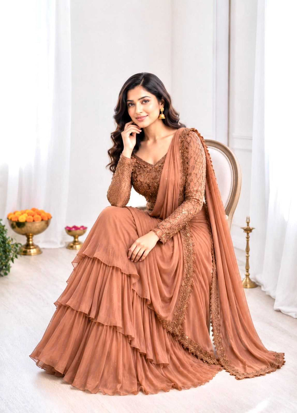 A woman seated on a wooden chair wearing a terracotta-colored traditional gown with a matching draped scarf, set in a bright, minimalist indoor room with white curtains and decorative gold accents.