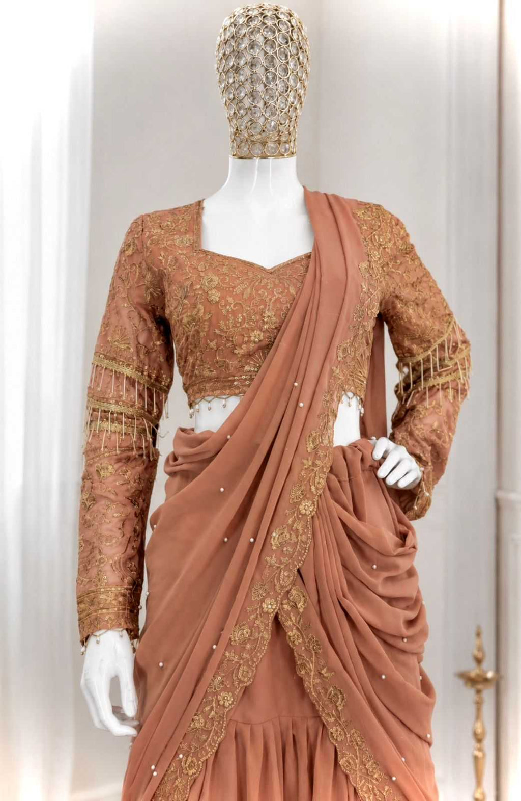 A peach-colored traditional Indian salwar suit set displayed on a mannequin. The set includes a long-sleeved kurta with gold embroidery and fringe, a matching draped sari with gold border detailing, and a beaded headpiece.