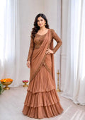 A woman models a traditional Indian salwar kameez outfit in a warm terracotta brown color, featuring a heavily embroidered long-sleeve kameez top and a tiered draped skirt, set against a soft white curtain background.