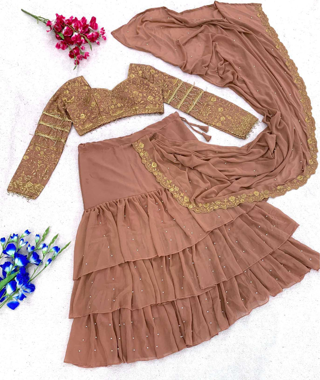 A women's traditional outfit laid flat on a white textured background, featuring a brown tiered skirt with gold embroidery, a matching long-sleeve blouse, and a draped dupatta. Decorative flowers are placed in the corners.
