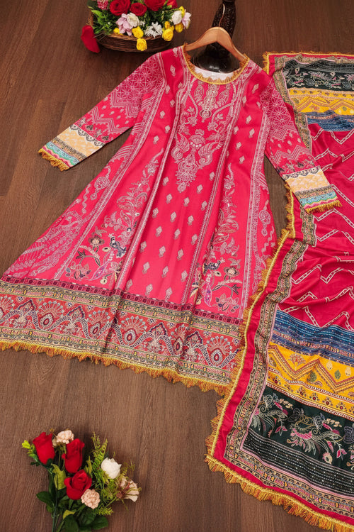 A bright pink traditional South Asian outfit set laid flat on a wooden floor, featuring a long kurta with intricate multicolor embroidery, a matching patterned dupatta, and decorative floral props at the corners.