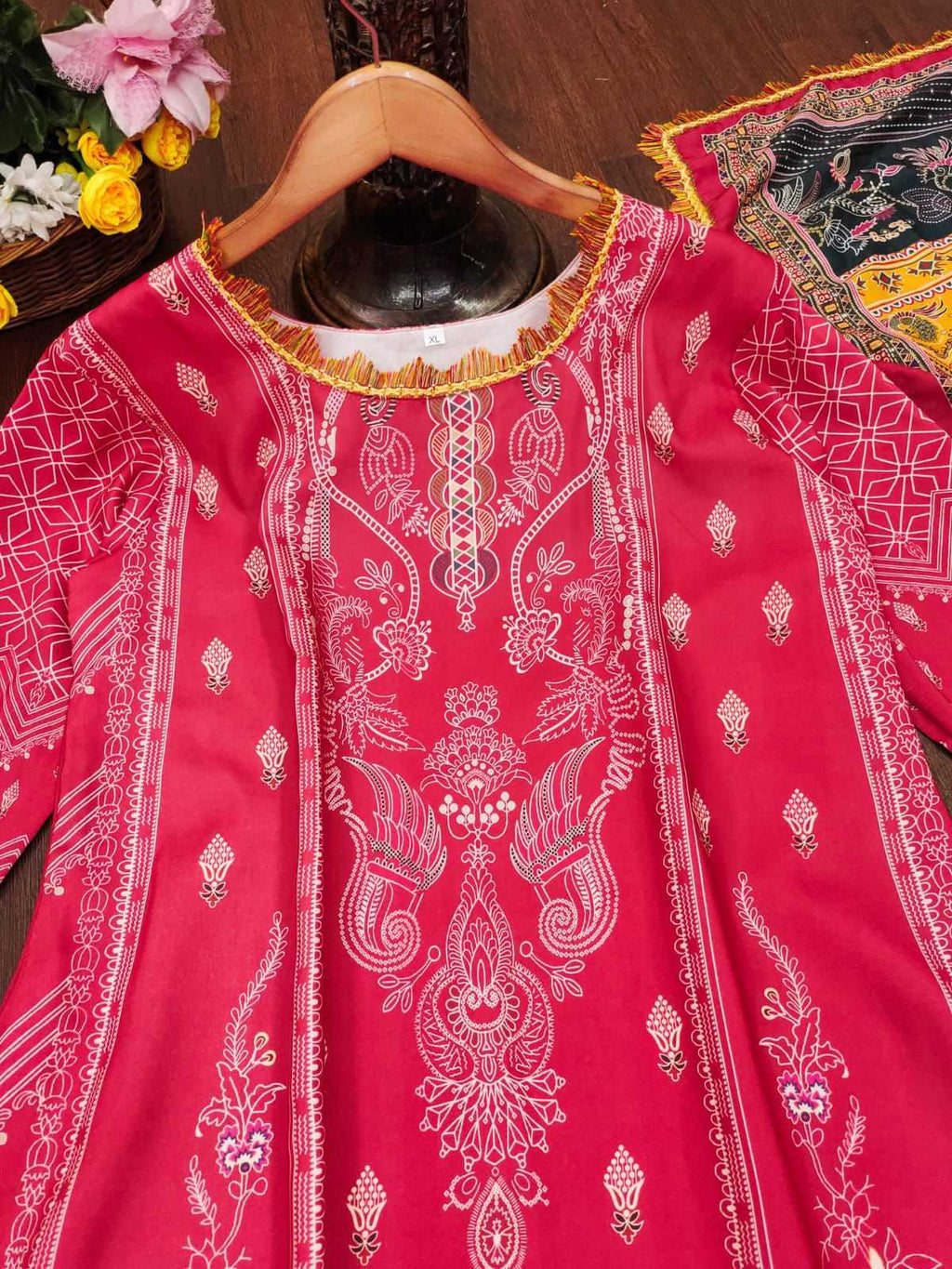 A bright pink embroidered kurta with intricate white and gold patterns, displayed on a wooden hanger against a dark wood background.
