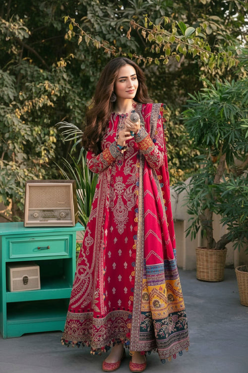 Woman in a vibrant red and patterned traditional outfit standing outdoors with greenery in the background.
