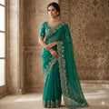 Woman wearing a teal saree with intricate embroidery in an elegant room.