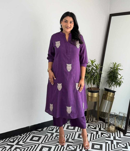 Woman wearing a purple dress with white patterns standing in a room with a mirror and plants.