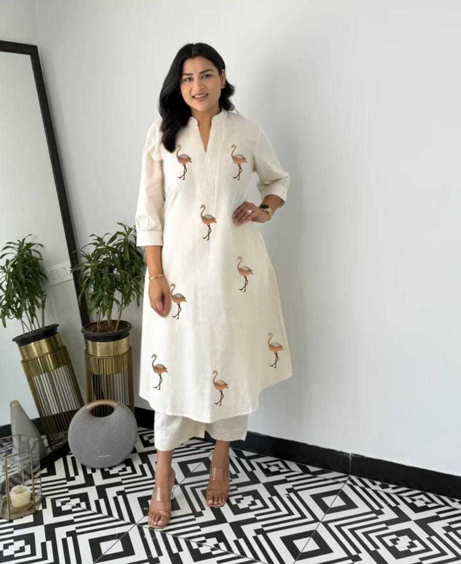 Woman wearing a white dress with flamingo patterns in a room with plants and decorative items.