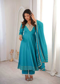 A woman models a teal blue traditional Indian salwar kameez suit with a matching dupatta. The outfit features intricate gold embroidery on the neckline and borders, paired with matching trousers and a draped dupatta.