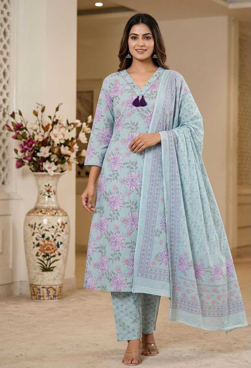 A woman models a light blue three-piece traditional outfit with floral purple and green prints, featuring a kurta, matching pants, and a dupatta, set against an elegant indoor background.