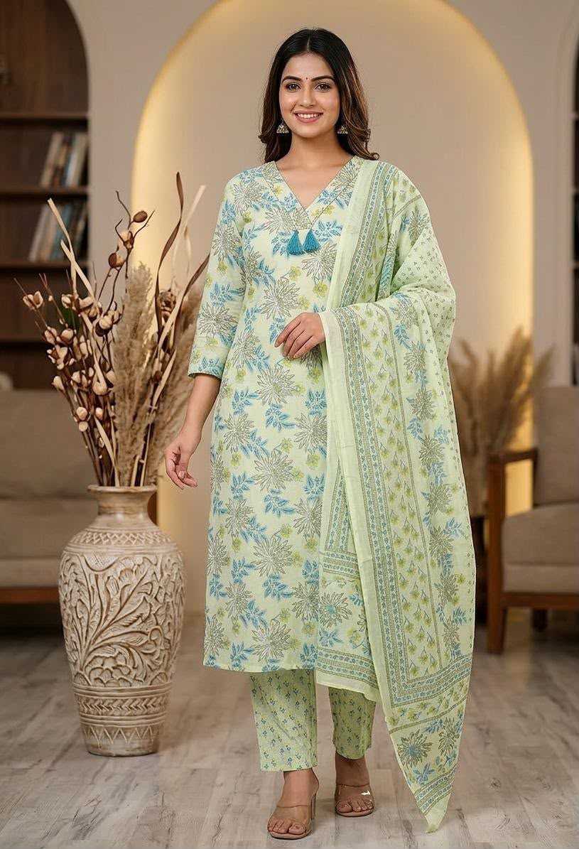 A woman modeling a light green and blue floral printed ethnic suit with a matching dupatta in a warm, modern living room setting.