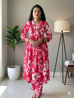 Woman wearing a red floral dress standing in a room with a plant and lamp.
