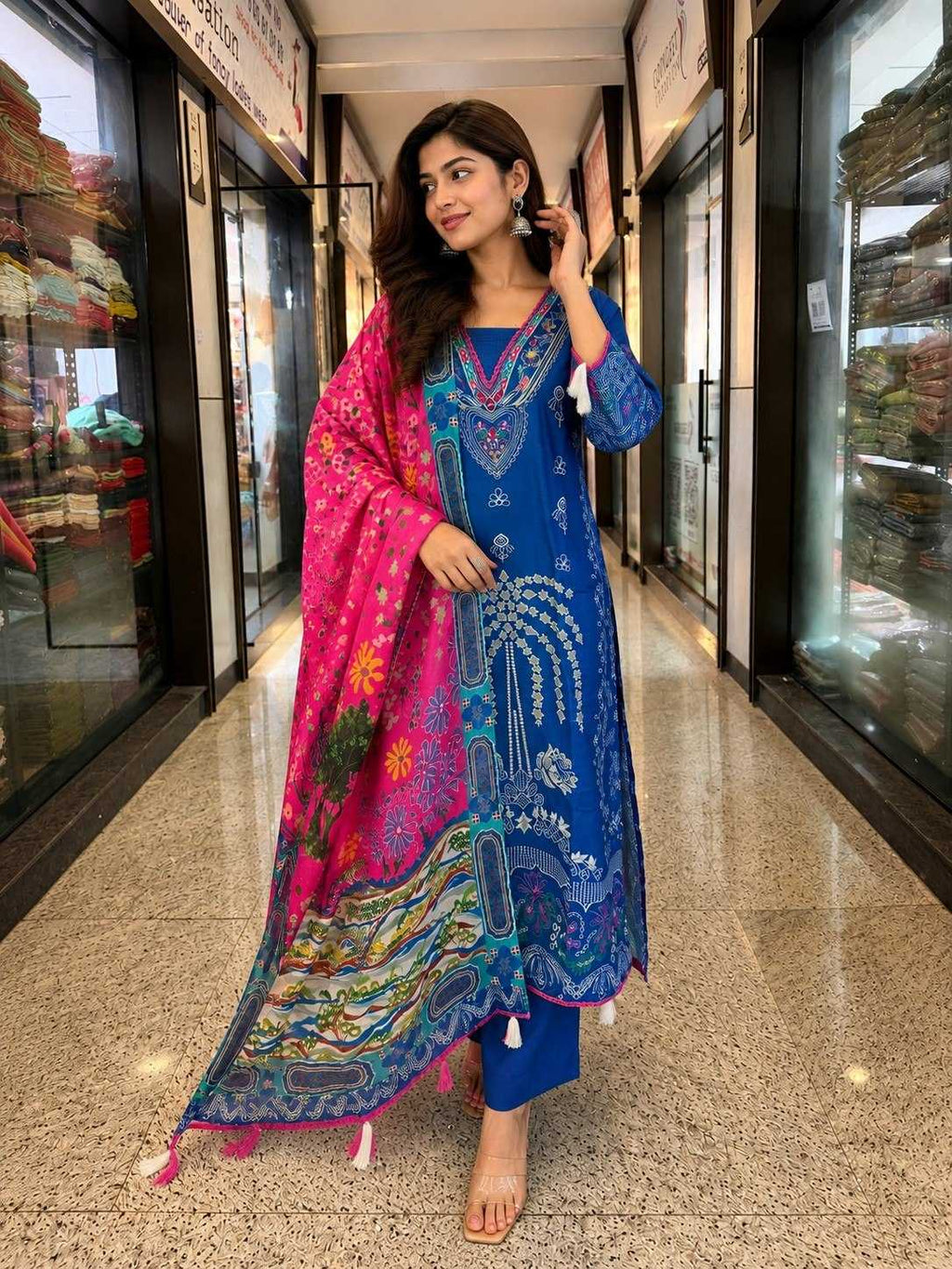A woman models a traditional South Asian outfit consisting of a royal blue kurta with white embroidery, matching blue trousers, and a vibrant pink dupatta with multicolor paisley patterns, standing in a commercial hallway.