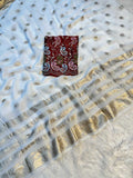 A white georgette saree with metallic gold foil dot patterns, a wide gold zari border, and a small folded red fabric patch with colorful paisley embroidery.