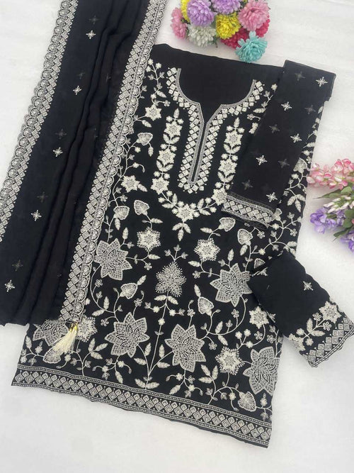 Black cotton three-piece salwar suit set with intricate silver floral embroidery, featuring a kurta, matching trousers, and a dupatta with silver borders and star motifs, styled on a white surface with decorative flowers.