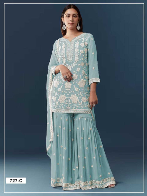 A woman modeling a light blue traditional three-piece outfit with white floral embroidery, featuring a long kurta, flared pants, and a matching dupatta against a dark blue studio background.