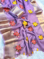 A lavender purple saree with metallic gold borders and embroidered floral motifs in red, yellow, and green, draped on a white textured background.