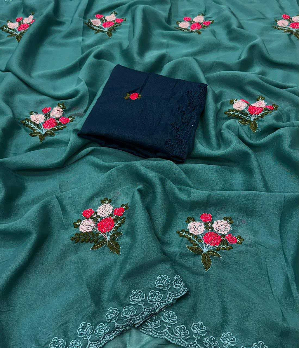 A teal green embroidered fabric, likely a salwar kameez or kurta fabric, draped to show its texture and design. The fabric features embroidered floral motifs in pink, white, and green, and has a folded dark navy blue fabric piece with matching embroidery placed in the center.