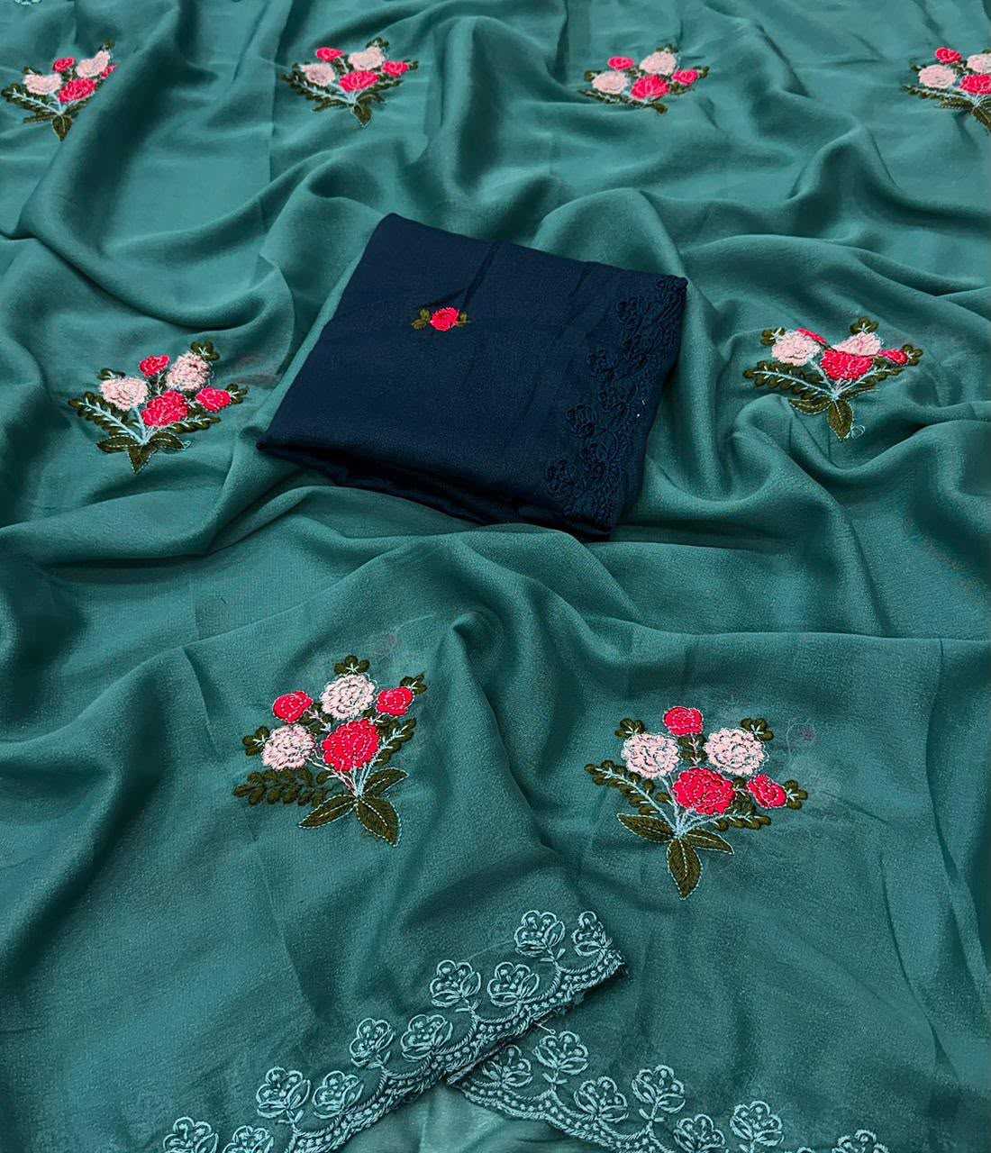 A teal green embroidered fabric, likely a salwar kameez or kurta fabric, draped to show its texture and design. The fabric features embroidered floral motifs in pink, white, and green, and has a folded dark navy blue fabric piece with matching embroidery placed in the center.