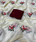 White silk-like fabric with embroidered floral motifs in red, pink, and green, draped to show its texture and a decorative beige lace border, with a folded dark red fabric piece placed in the center.