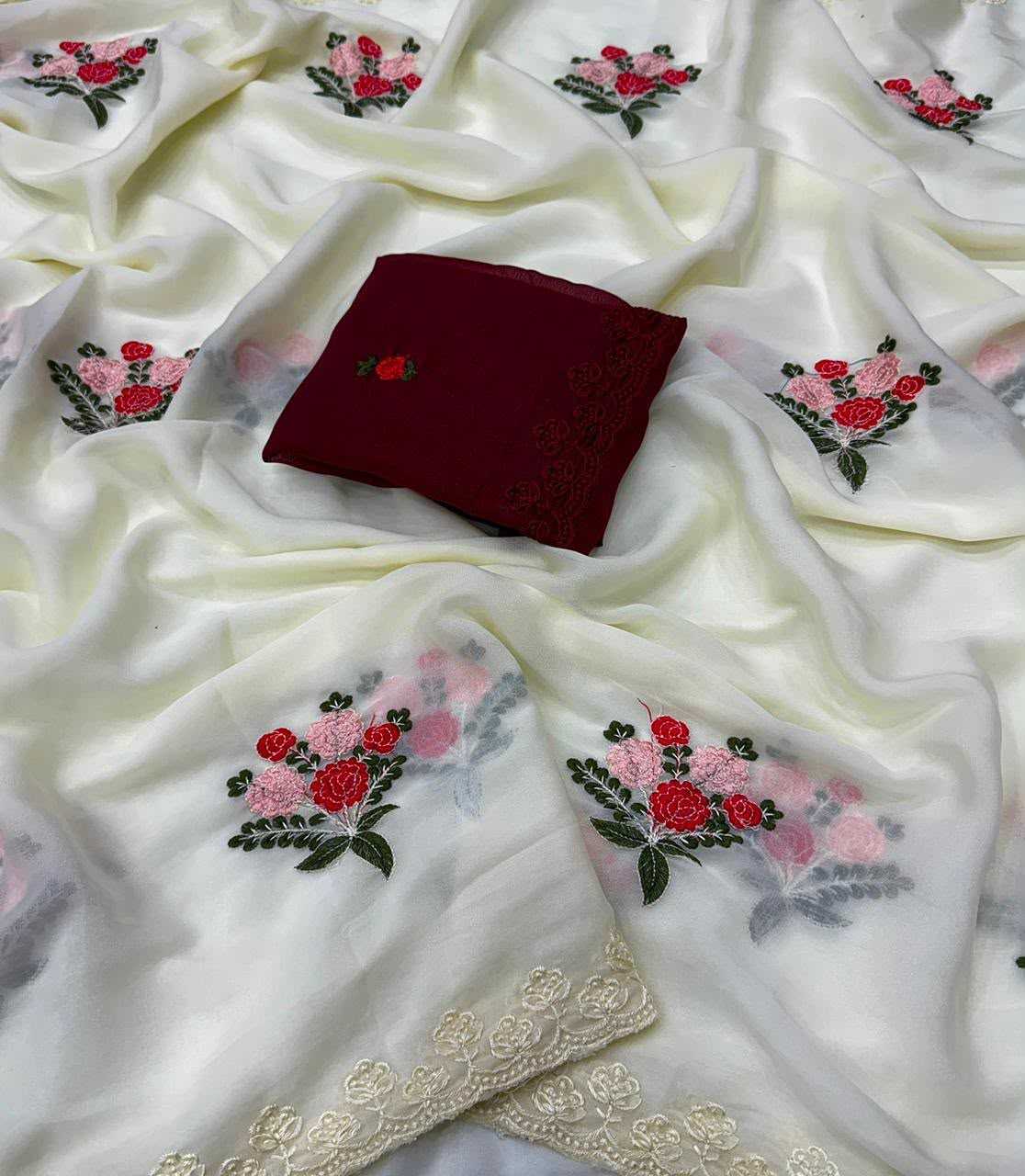 White silk-like fabric with embroidered floral motifs in red, pink, and green, draped to show its texture and a decorative beige lace border, with a folded dark red fabric piece placed in the center.