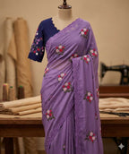A lavender purple saree with floral embroidery and a navy blue sleeveless blouse, displayed on a mannequin in a sewing room setting.