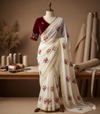 A cream-colored saree with pink floral embroidery and a deep maroon velvet sleeve, displayed on a mannequin in a warm, rustic sewing room setting.