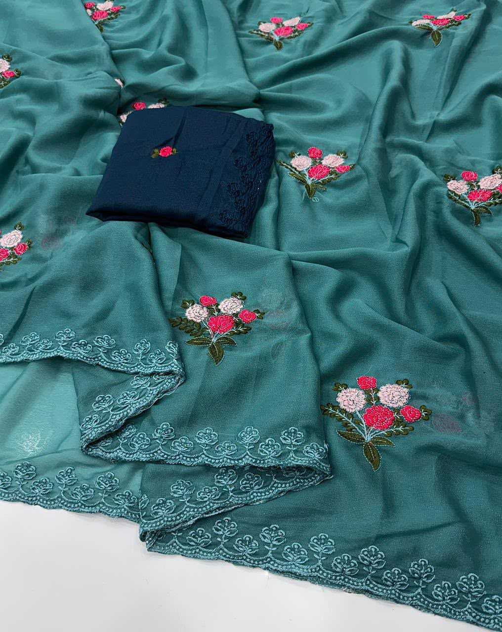 A teal green embroidered fabric, likely a saree or dupatta, draped to show its floral embroidery and scalloped lace borders. A folded dark navy blue fabric piece with a small matching floral embroidery is placed on top.