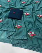 A teal green embroidered fabric, likely a saree or dupatta, draped to show its floral embroidery and scalloped lace borders. A folded dark navy blue fabric piece with a small matching floral embroidery is placed on top.