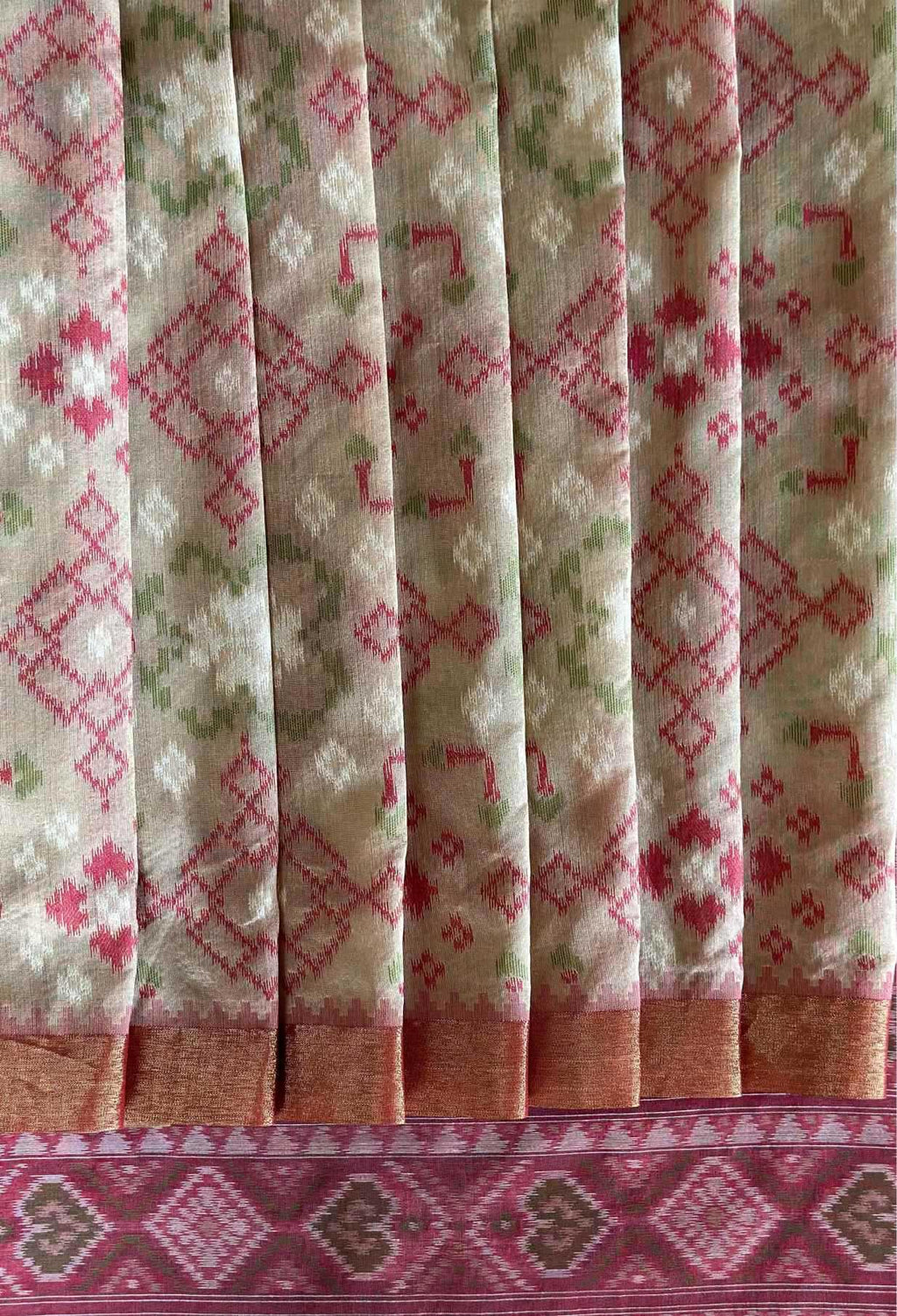 A close-up of a patterned fabric, likely a saree, featuring vertical pleats with a beige base, pink and green woven motifs, and a wide pink border at the bottom.