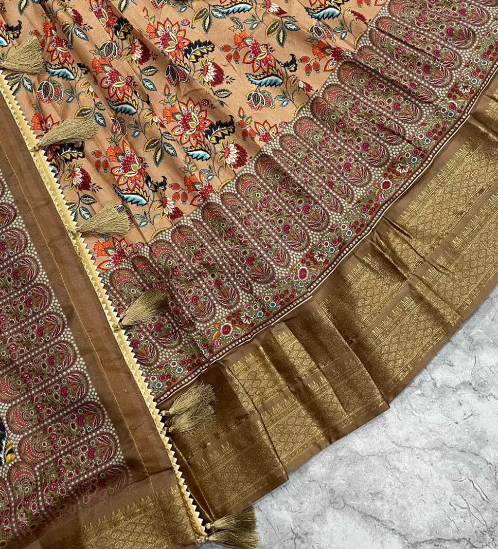 A close-up of a traditional Indian saree or fabric panel with a peach-beige base, dense floral and bird motifs in red, blue, green, and black, a wide metallic gold border with scalloped paisley patterns, and tassel trim, laid over a light marble surface.