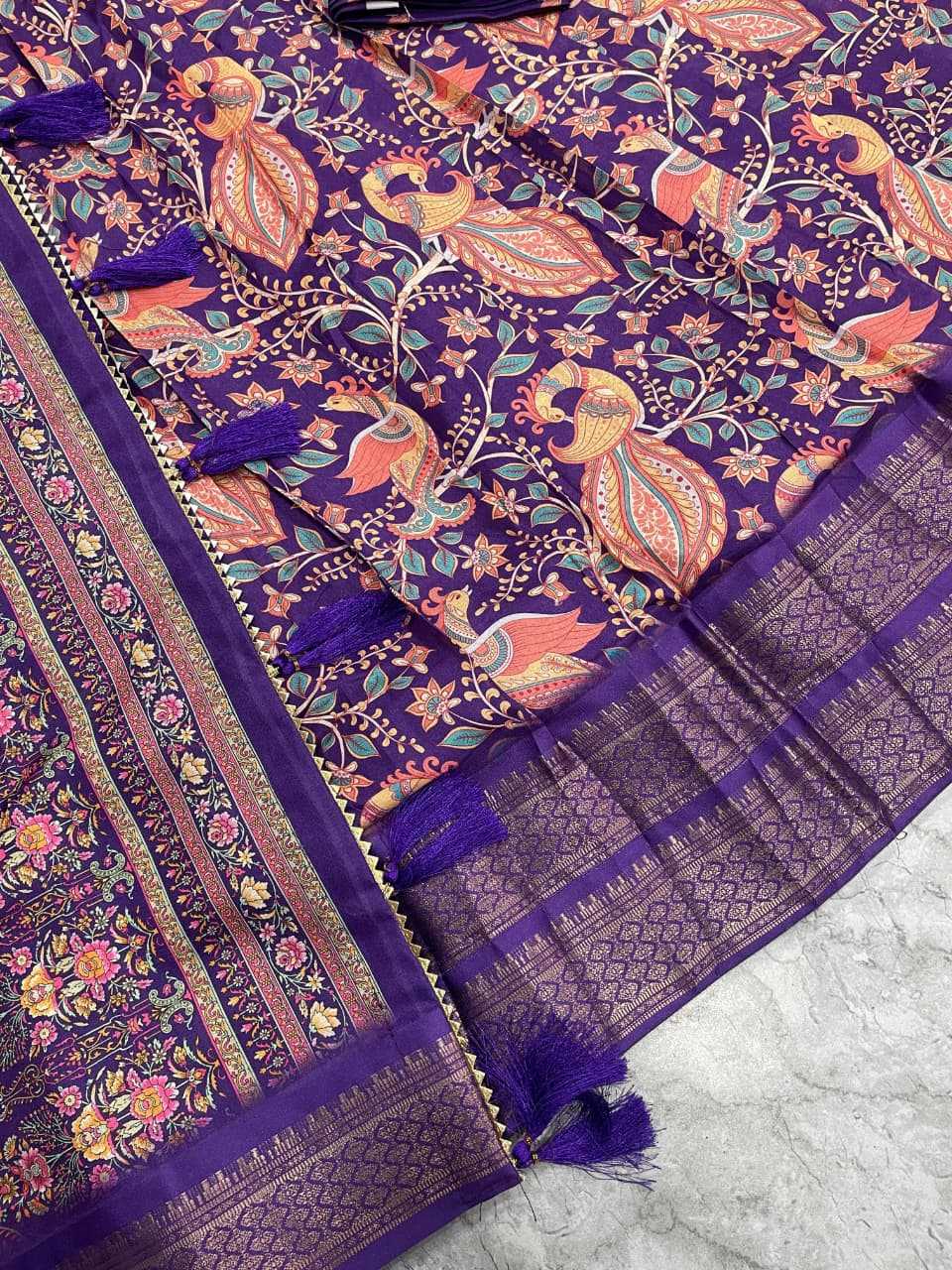A close-up of a purple printed fabric with ornate floral and bird motifs in orange, pink, and green, plus matching purple tassels and a patterned border.