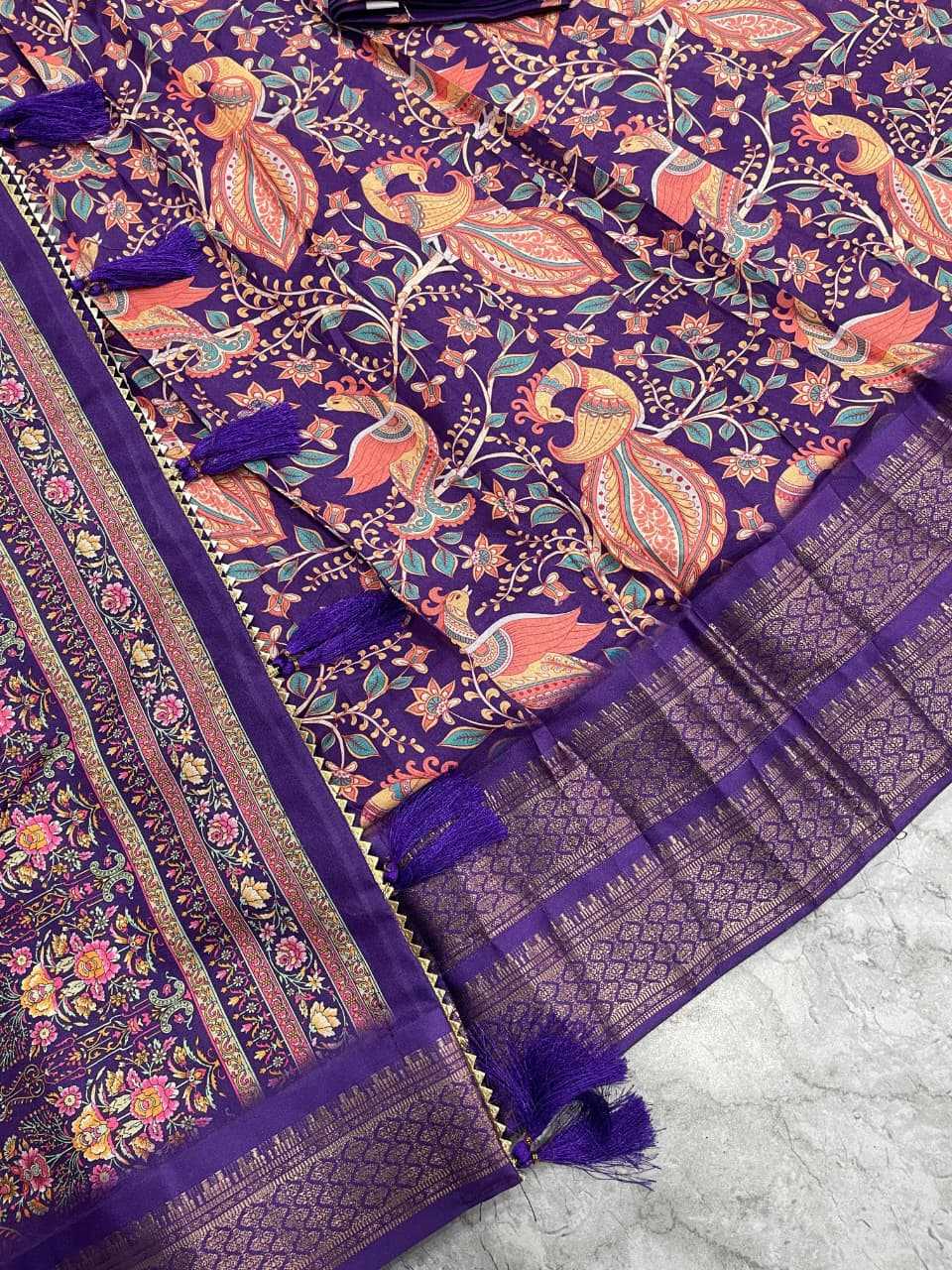 A close-up of a purple printed fabric with ornate floral and bird motifs in orange, pink, and green, plus matching purple tassels and a patterned border.
