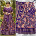 A purple and pink printed traditional Indian skirt set with a matching blouse, shown both worn by a model outdoors and laid flat. The fabric features intricate floral and peacock motifs with metallic zari borders.