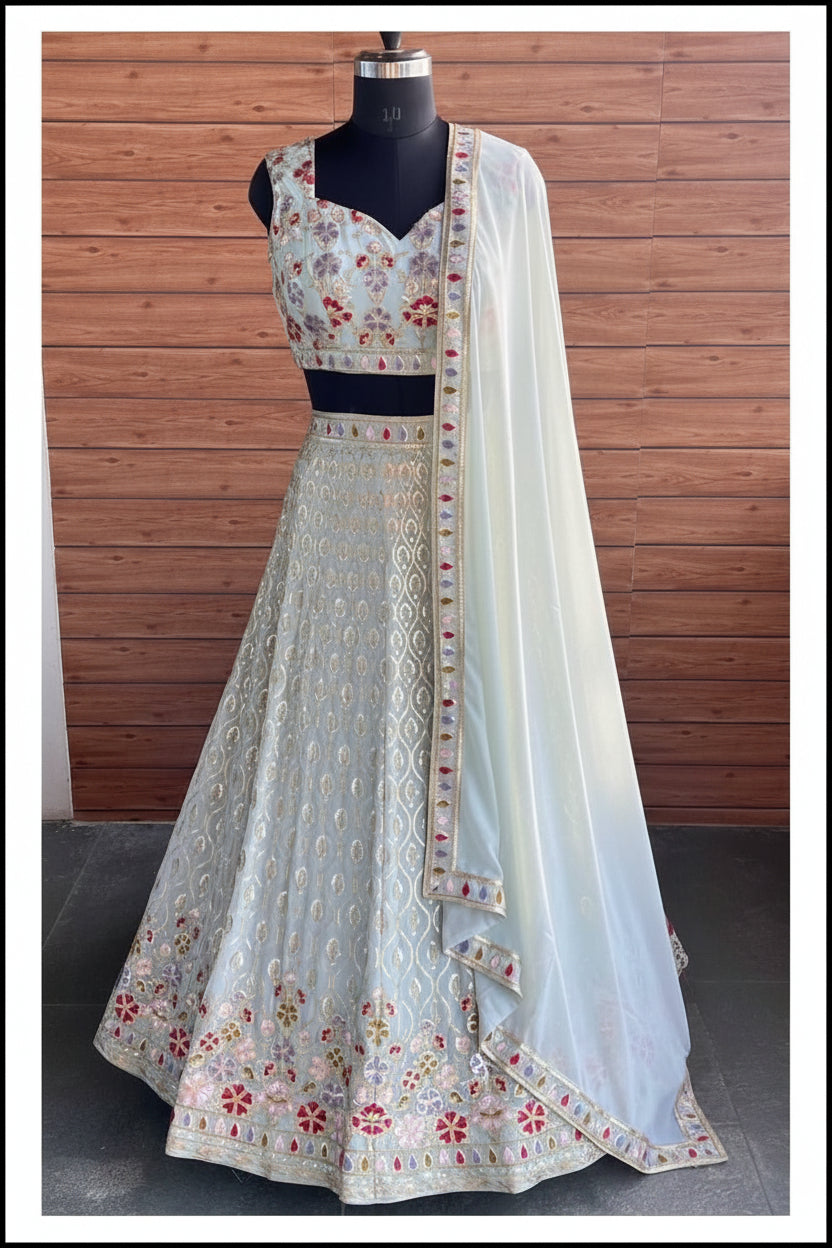 A sleeveless light blue lehenga choli set with floral embroidery and a matching dupatta draped over a black mannequin, set against a wooden wall background.