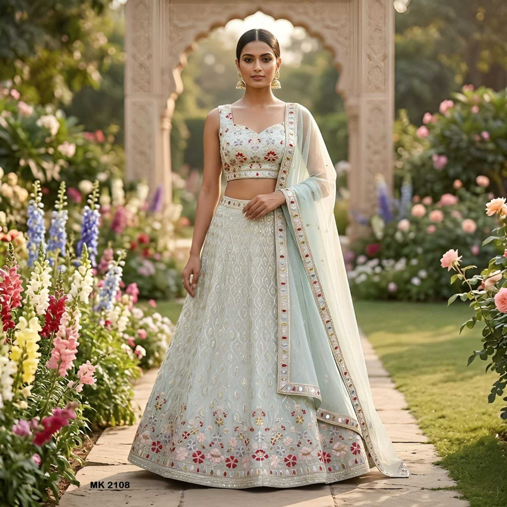 A woman stands in a garden wearing a light blue and white embroidered traditional outfit with a matching sheer dupatta. The outfit features intricate floral embroidery and a flared skirt, with a stone archway and colorful flowers in the background.