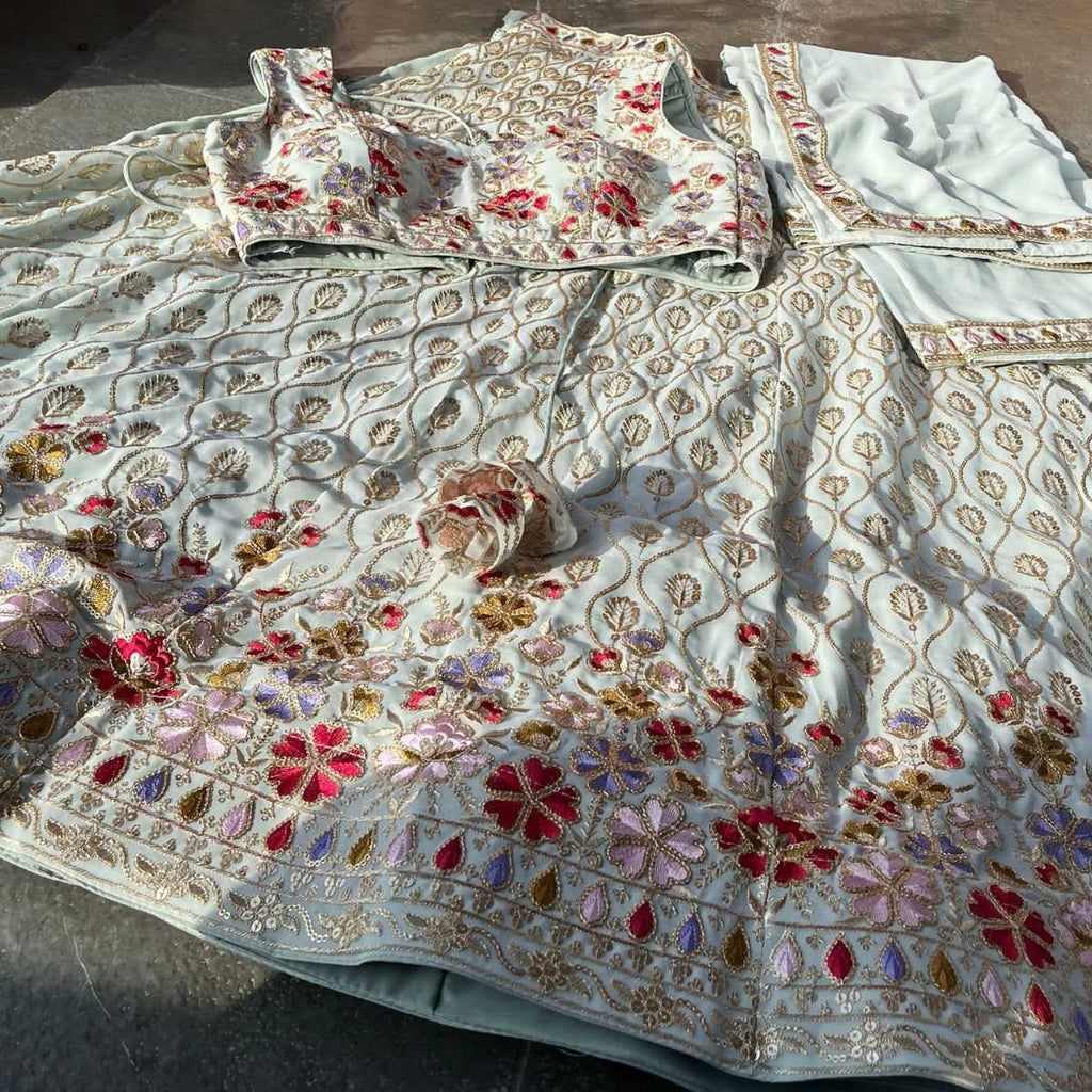 An embroidered off-white ethnic outfit laid out on a dark floor, featuring a printed and heavily embellished floral lehenga skirt, a matching printed blouse, and a coordinating dupatta.