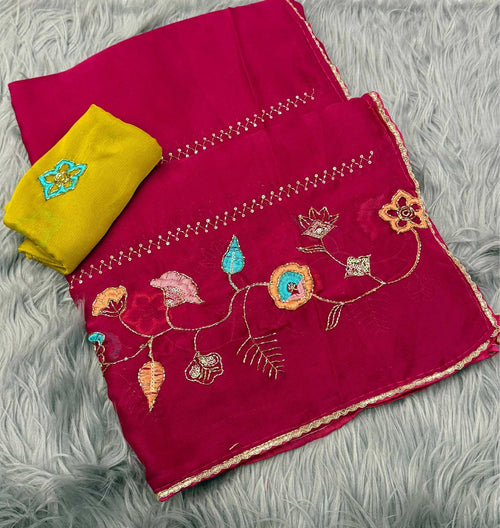 A folded magenta pink fabric piece with colorful floral embroidery and gold border trim, shown alongside a folded yellow fabric piece on a gray faux fur background.