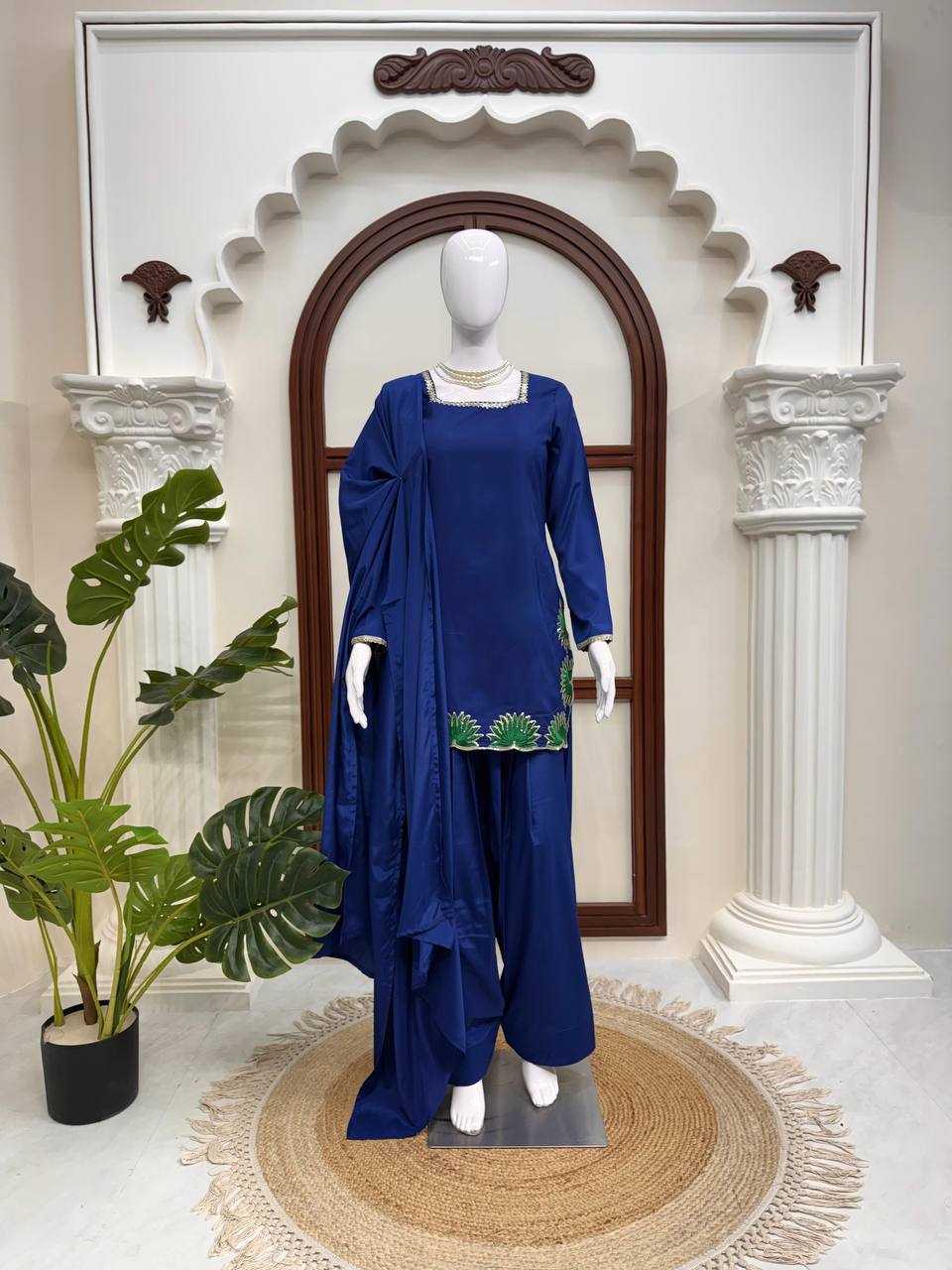 Mannequin wearing a blue outfit with green embroidery in front of a decorative archway.