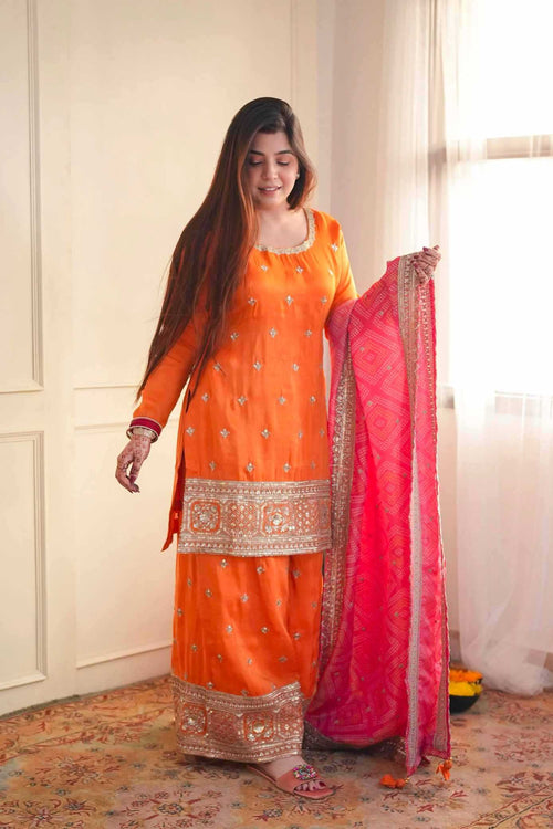 A woman modeling an orange traditional ethnic outfit with silver embroidery, holding a bright pink dupatta with gold borders in an indoor setting with cream walls and a patterned rug.