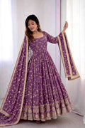 A woman modeling a purple Anarkali gown with gold floral embroidery and a matching dupatta with gold borders, set against a white curtain background.