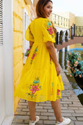 Woman wearing a yellow dress with floral embroidery in an outdoor setting.
